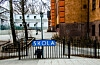 Blå «SKOLA»-skilt på en port foran et inngjerdet skoleområde med bygninger og trær.
