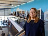 Kvinne står i et moderne kontoratrium med glasrekkverk og blå vegg i bakgrunnen.