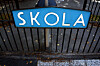 Blått skilt med teksten «SKOLA» på svart metallgjerde.