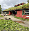 Rød bygning med torvtak, piknikbord og grønt område foran.
