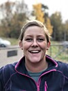 Smilende kvinne i mørk jakke står utendørs med trær og bygninger i bakgrunnen.
