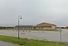 Lavt beige bygg med parkeringsplass, gress og gatelykt under en overskyet himmel.
