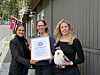 Tre kvinner viser fram et prisdiplom og en måke-bamse foran et barnehagebygg.