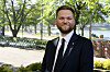 Dresskledd mann står foran trær, sykler og vann i et urbant utendørsmiljø.
