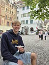 Ung mann i mørk hettegenser sitter i en brosteinslagt gate med historiske hus i bakgrunnen.