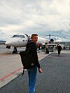 Reisende med ryggsekk på en flyplass foran et passasjerfly under en overskyet himmel.
