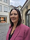 Smilende kvinne i rosa blazer foran butikker og historiske bygninger i en brosteinsgate.