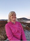 Inger Alsaker i rosa jakke står utendørs med fjell, bebyggelse og sjø i bakgrunnen.